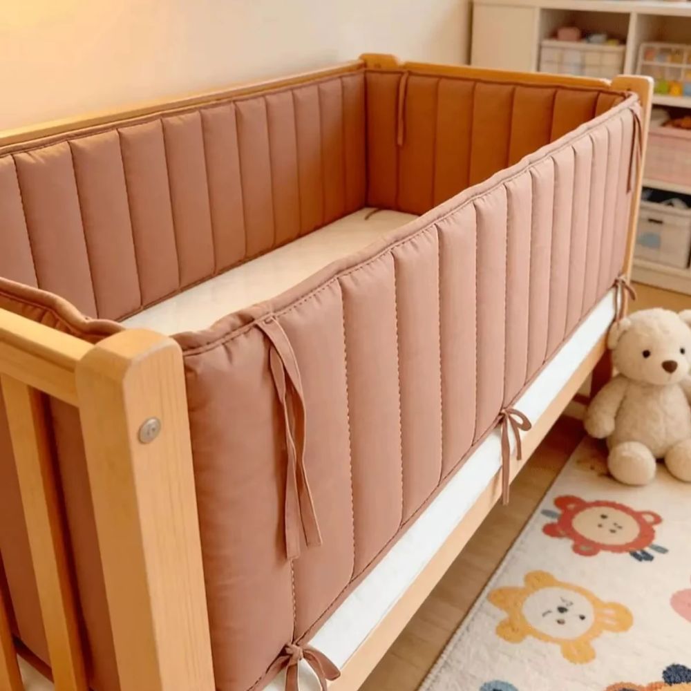 Terracotta cot bumper with vertical quilting tied to a timber cot, with teddy bears and a cartoon rug beside it