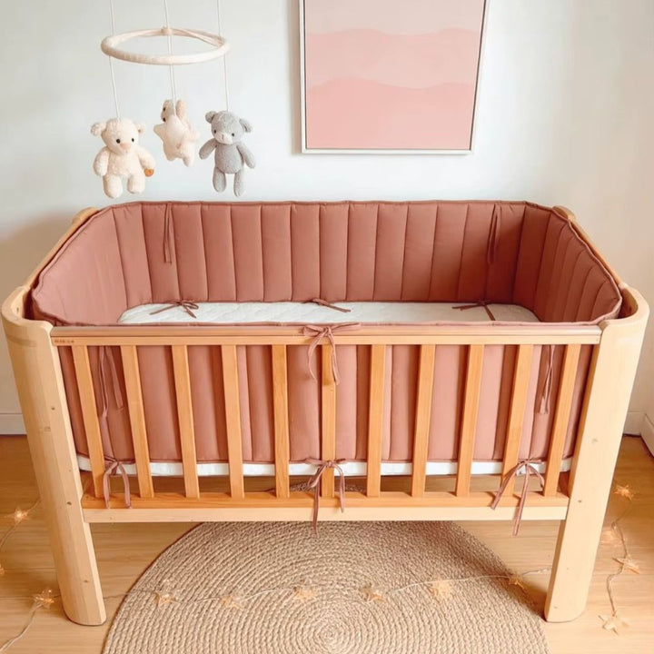 Terracotta quilted cot bumper tied around a timber cot, with a teddy bear mobile and jute rug in a soft pink nursery