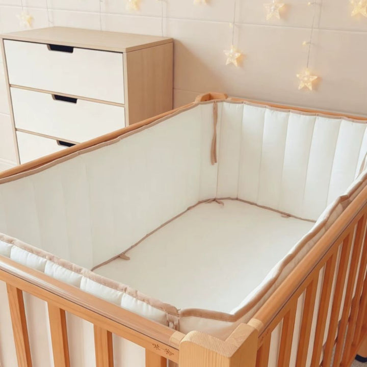 White cot bumper lining a timber cot in a neutral nursery with a chest of drawers and star fairy lights